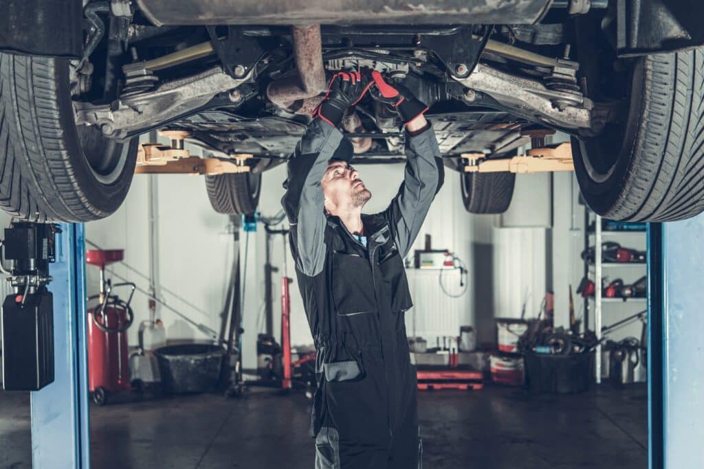 A motor trader is under a car, working on a repair in a garage. Demonstrating the need for service and repair garage insurance.